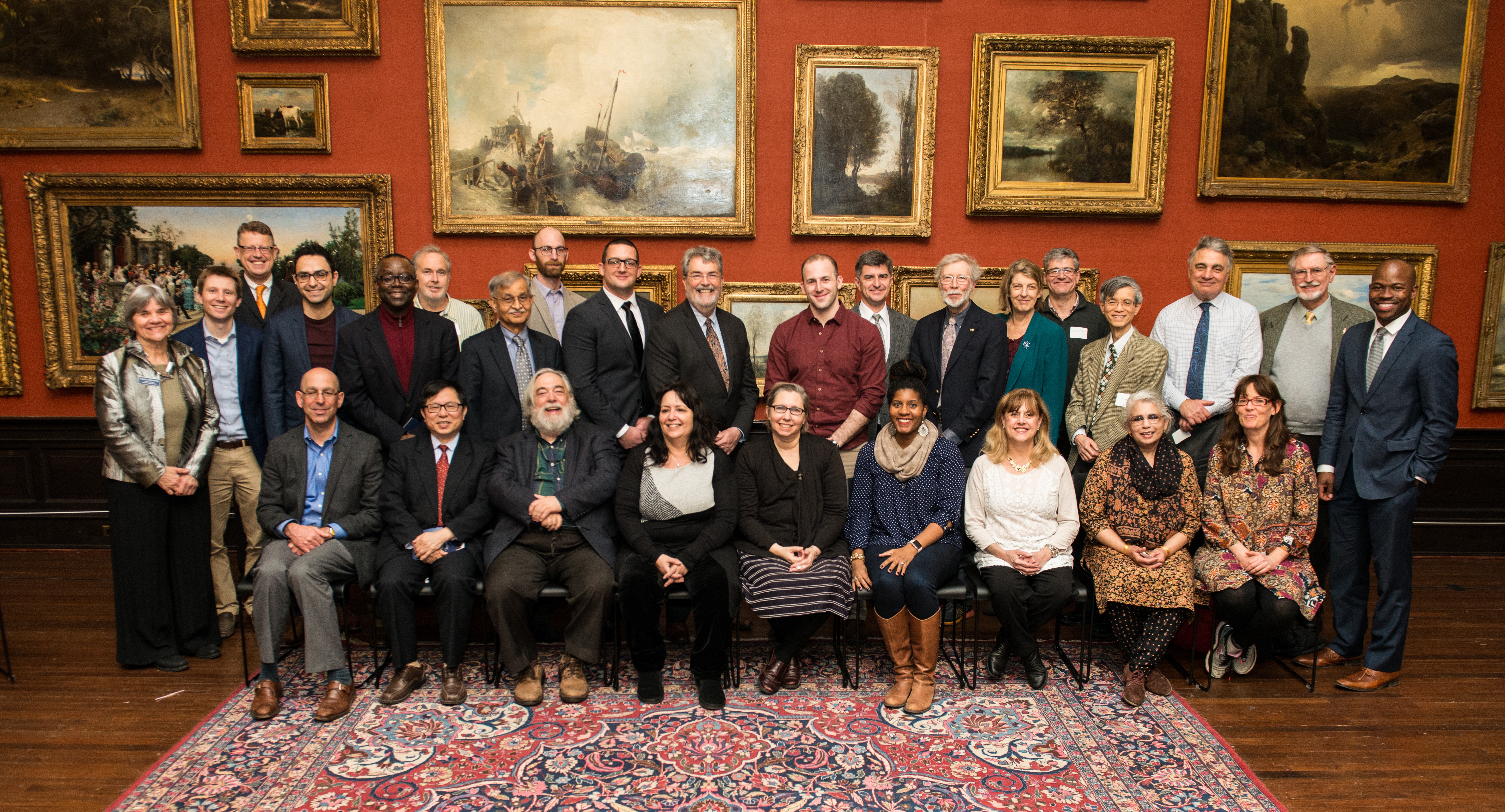 Men and women sit in two rows. They are posed in front of a wall with several large framed paintings.